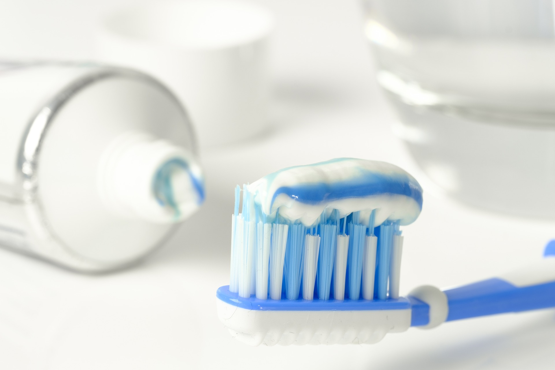 Close-up of a toothbrush with blue and white toothpaste on its bristles. In the background, theres a white toothpaste tube with a little paste on its opening and a clear glass filled with water.