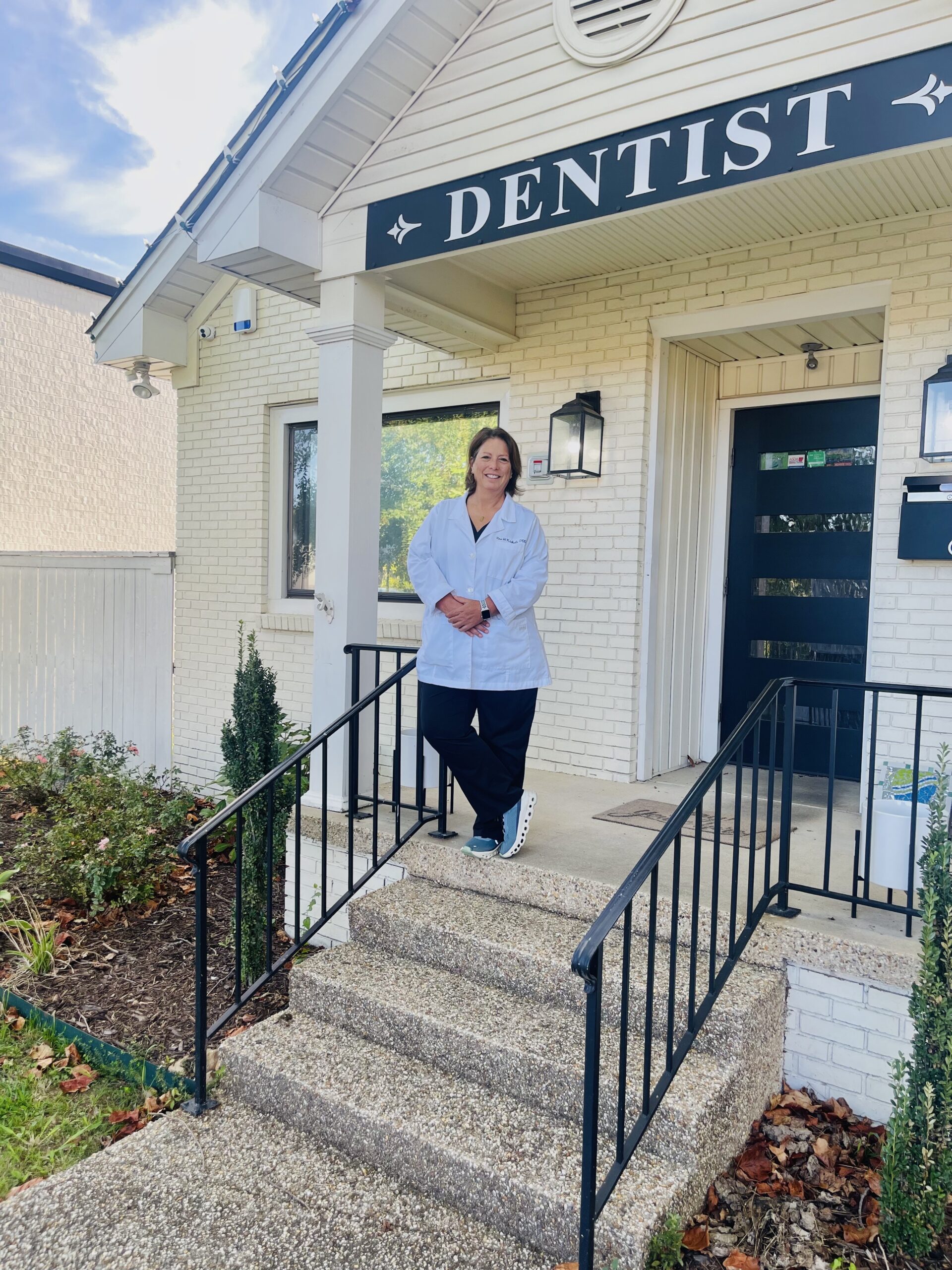 A dentist in a white coat stands smiling on the steps outside a building with a sign that reads DENTIST above the door. The building is white with black railings and has plants along the walkway.