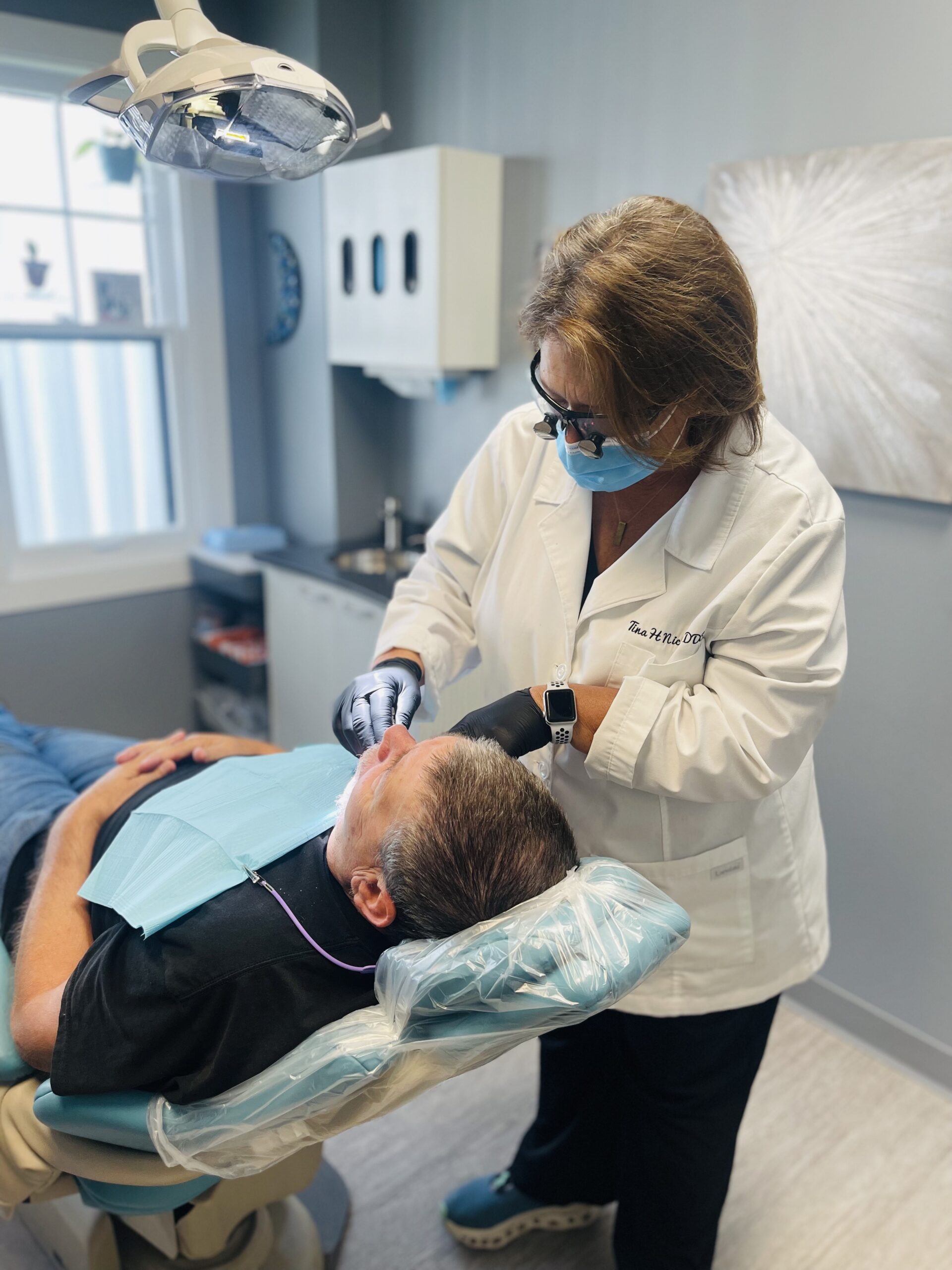 A dentist wearing a mask and gloves examines a patient lying in a dental chair in a bright, modern dental office. Dental tools and equipment are visible in the background.