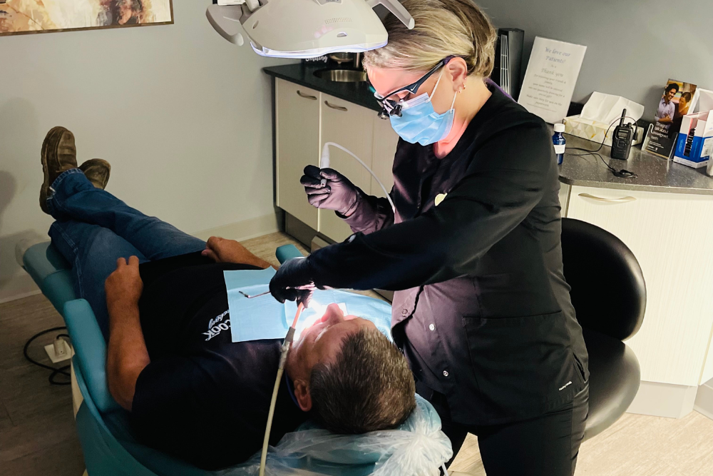 A dental professional wearing a mask and gloves works on a patient’s teeth in a dental clinic, with the patient lying back in the chair under a bright examination light.