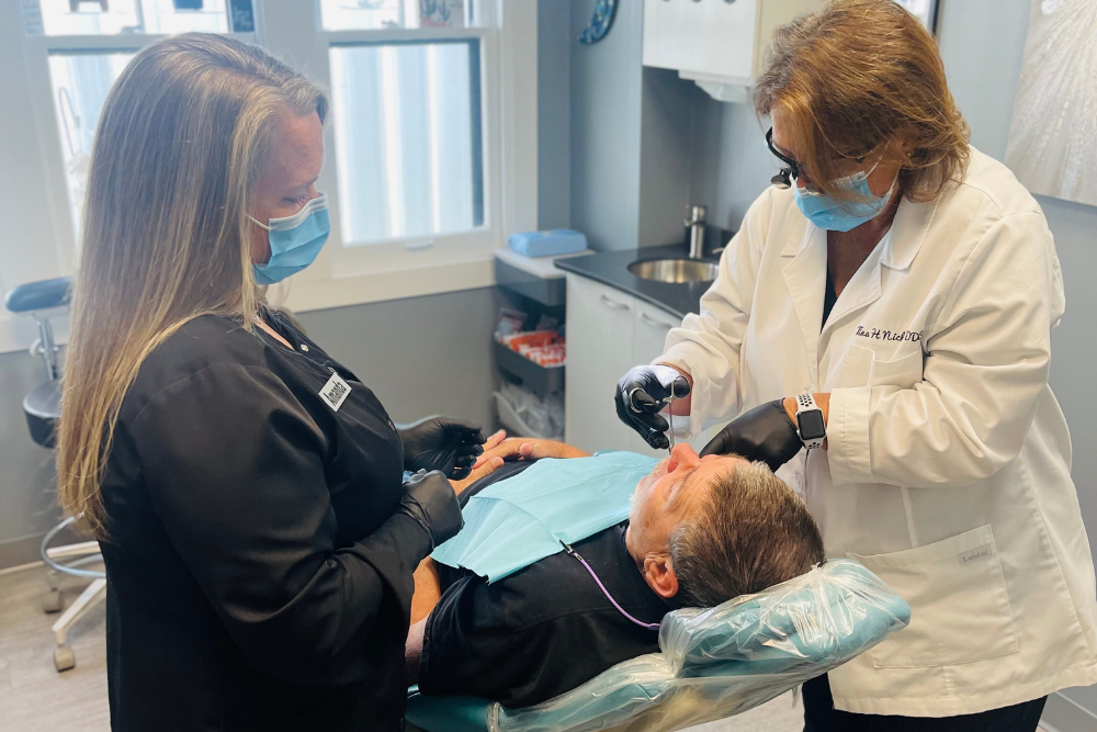 Two dental professionals in masks and gloves work on a male patient lying back in a dental chair inside a bright, modern dental office. One is using a tool on the patient while the other assists.