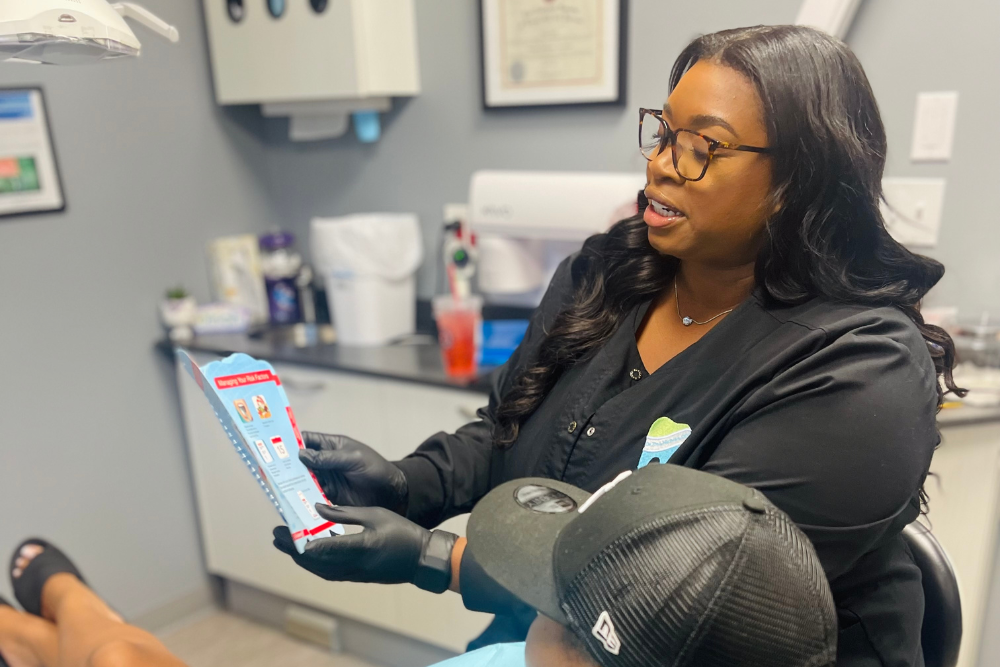 A dental professional wearing black gloves and glasses explains a brochure to a patient seated in a dental clinic. The patient is wearing a cap, and dental equipment is visible in the background.