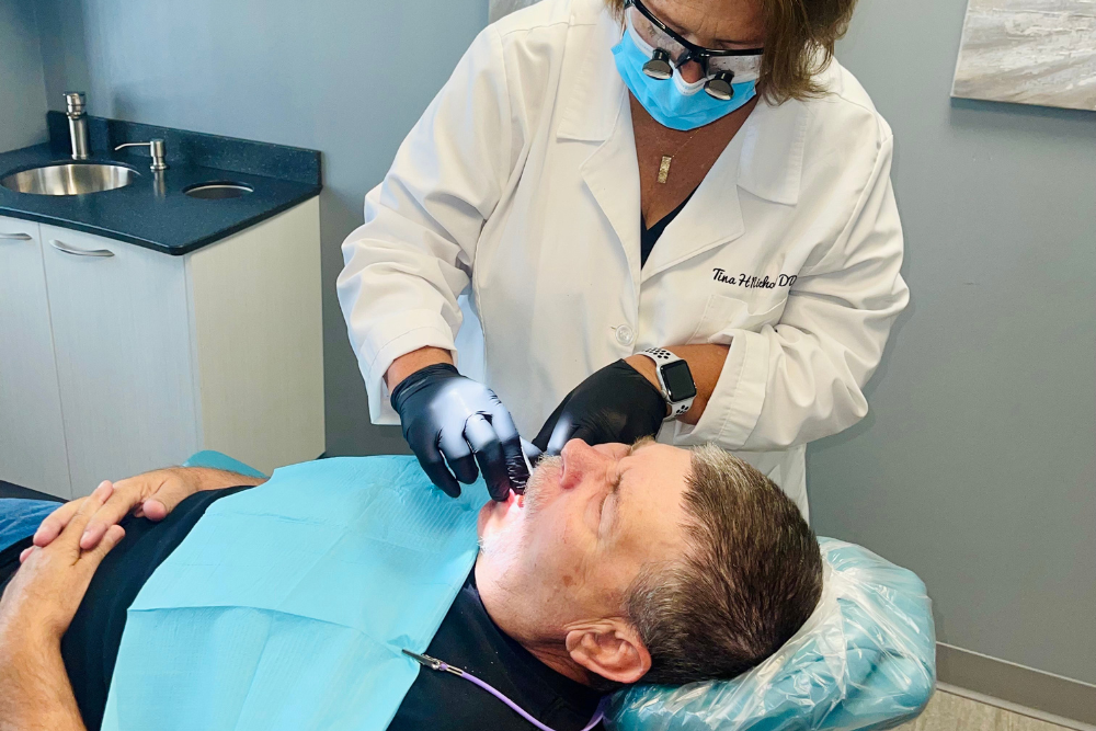 A dentist wearing gloves and a mask examines a patient’s mouth as he reclines in a dental chair, covered with a blue protective drape, in a modern dental office.